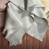 Sage textured 100% merino wool baby blanket with fringes on a brown surface, featuring the brand 'RIOJA'.