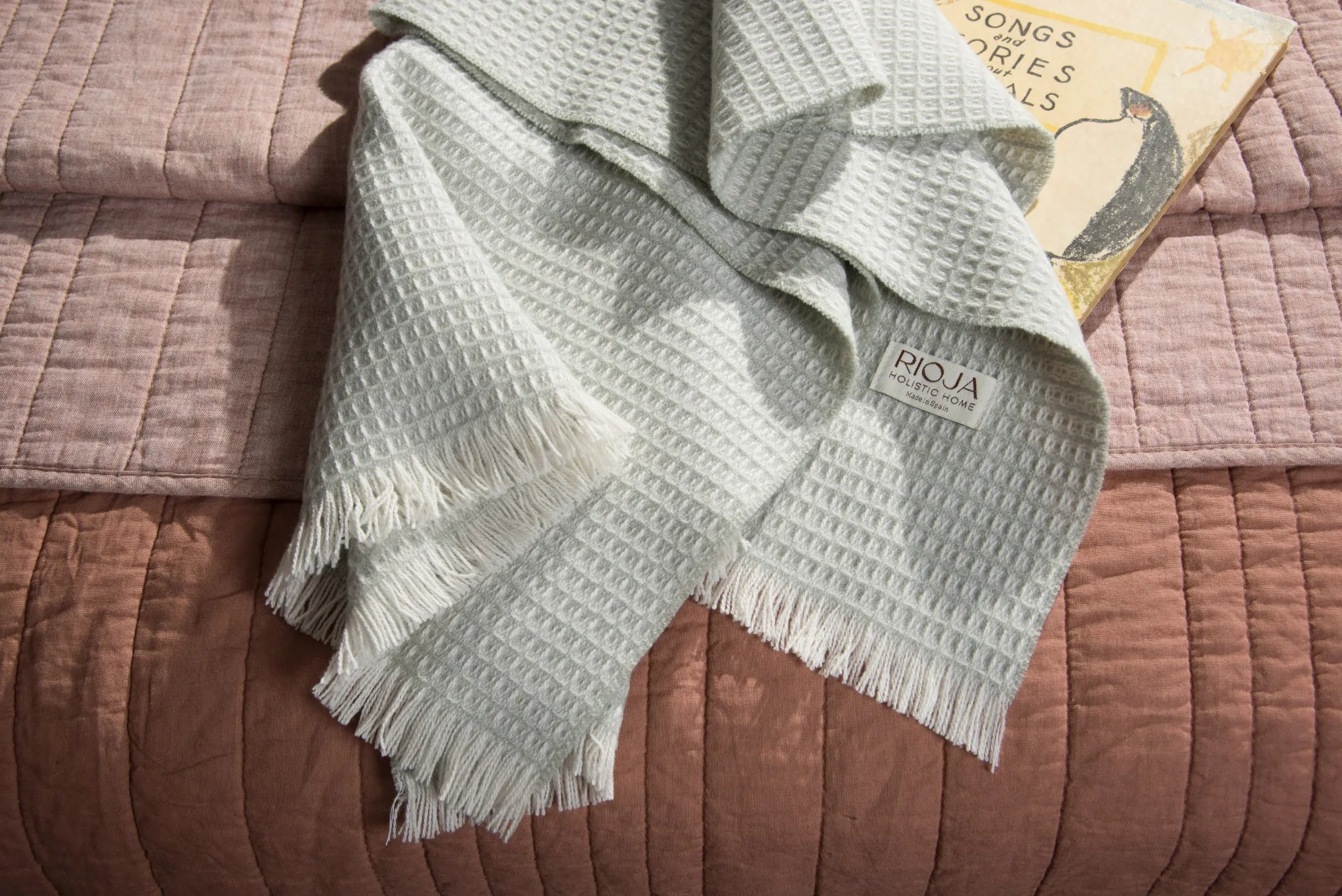 Sage textured 100% merino wool baby blanket with fringes on a brown surface, featuring the brand 'RIOJA'.