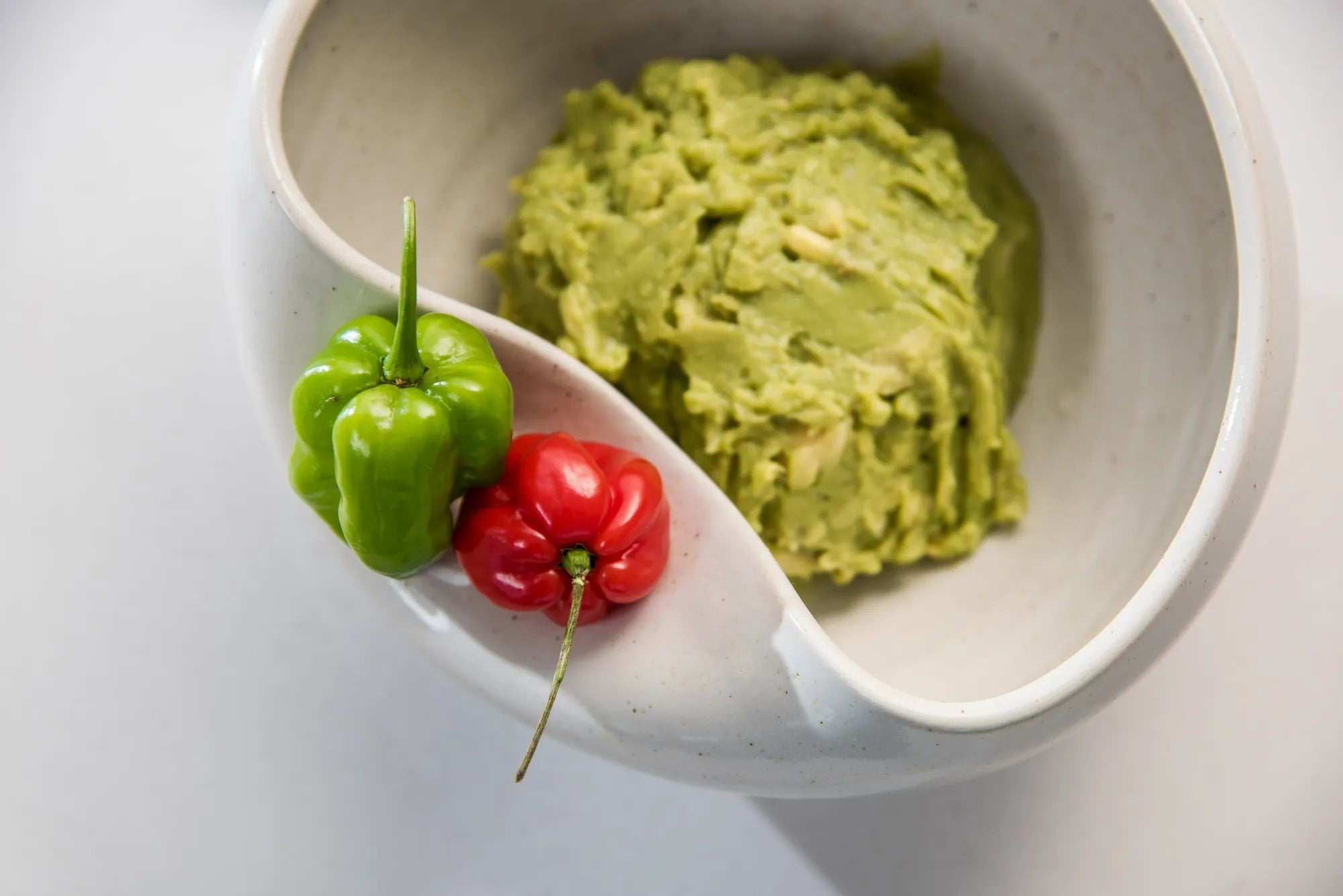 Guacamole in a ceramic Cuenko bowl with green and red peppers on a white tabletop