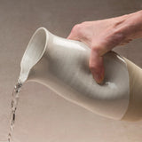 Hand pouring water from a ceramic Jarra pitcher on a neutral background