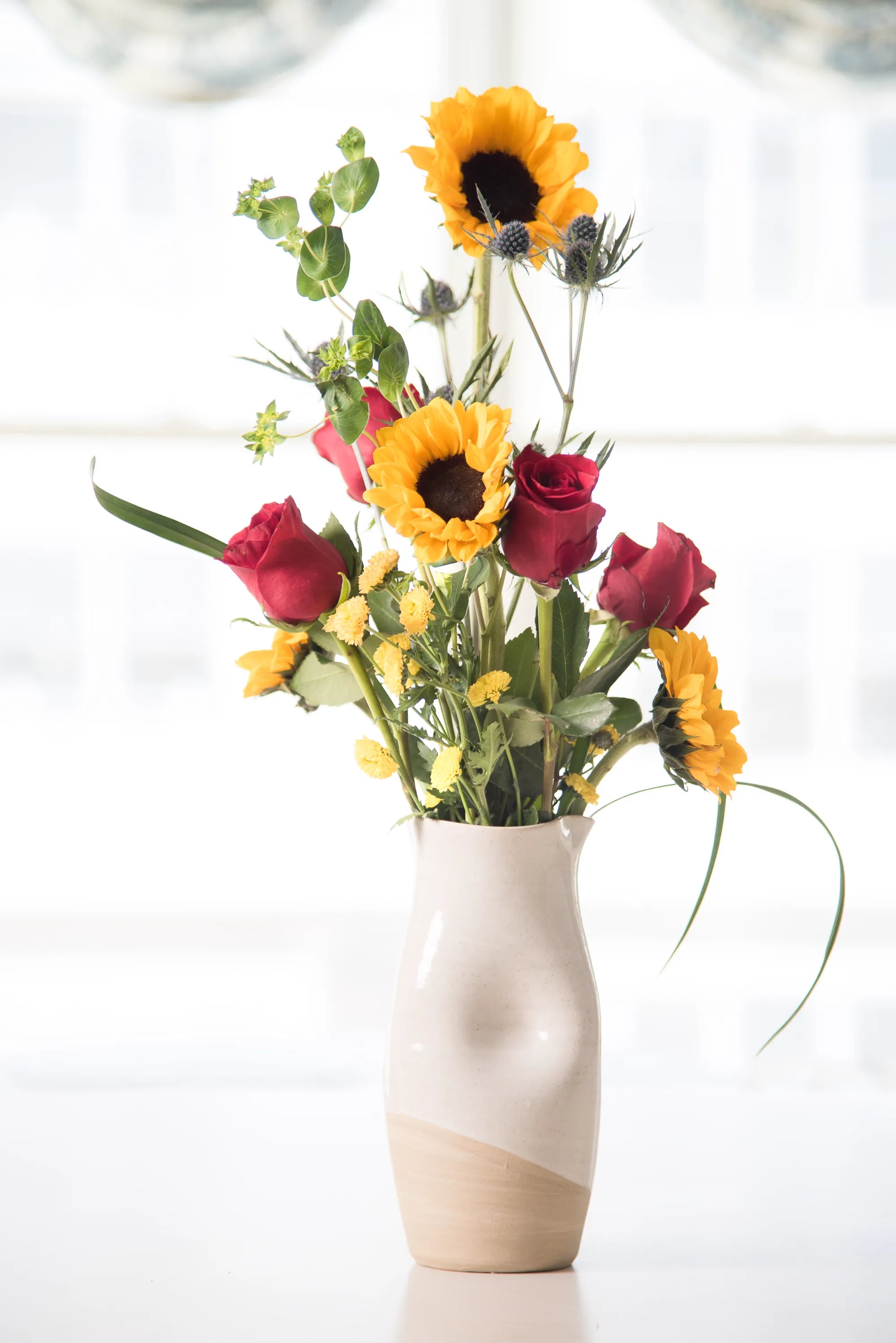 Floral arrangement with sunflowers and red roses in a Jarra pitcher vase on a light background
