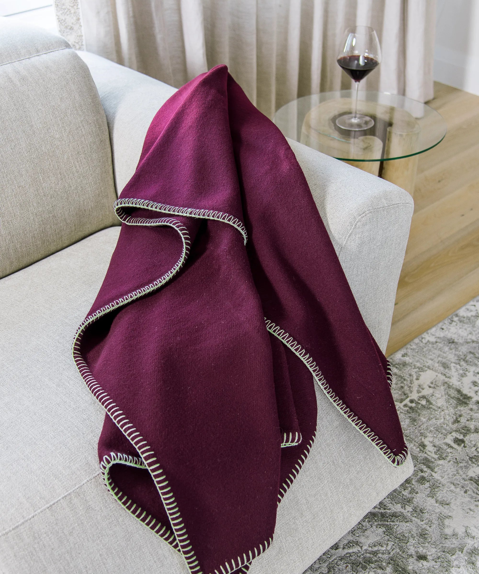 Plum 100% merino wool blanket draped over a white sofa with a glass table and wine glass in the background.