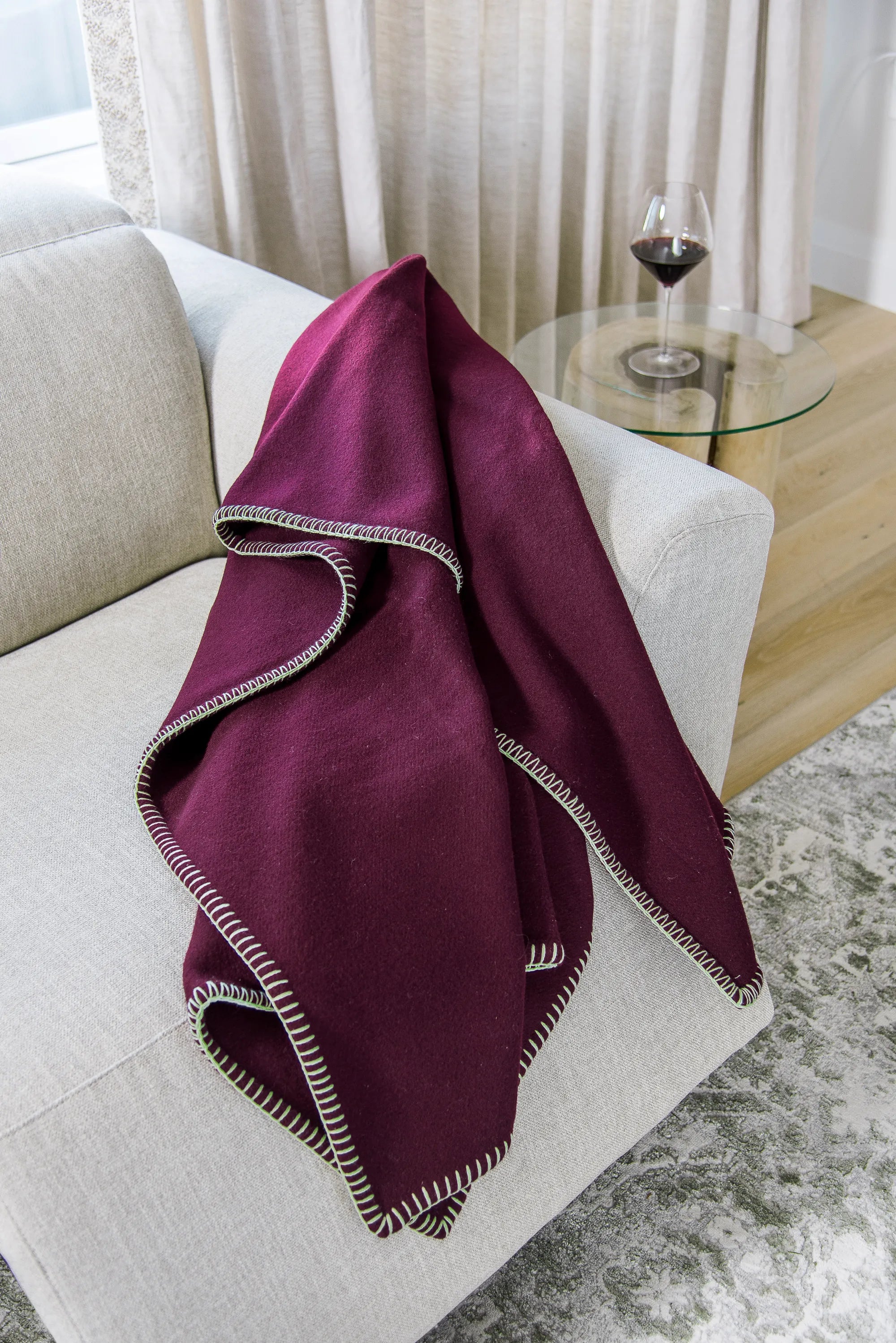 Plum 100% merino wool blanket draped over a white sofa with a glass table and wine glass in the background.